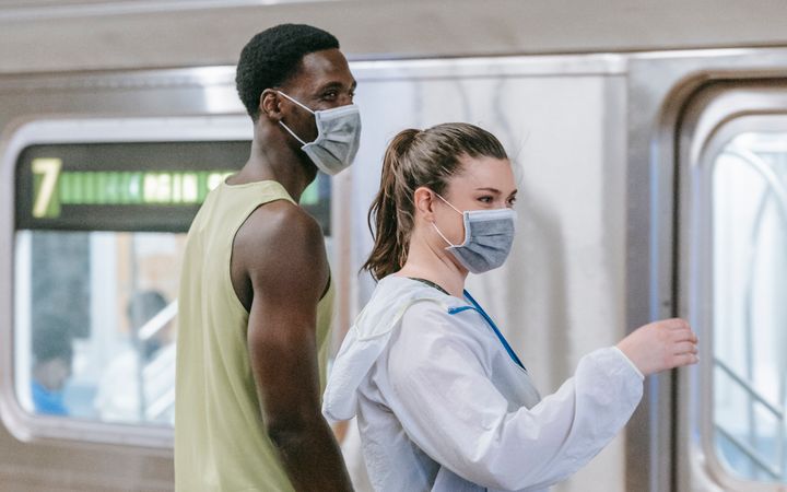 Two adults wearing masks stand at a train station. Captured in a vertical composition.