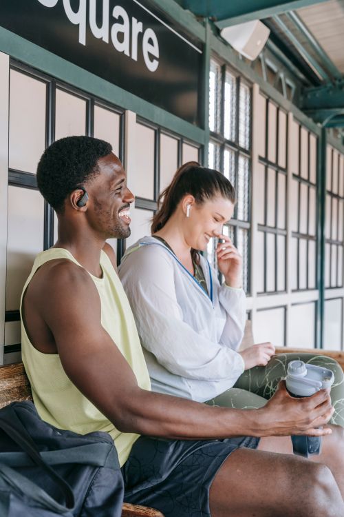 Free Two friends sitting on a bench, smiling and talking with earbuds, creating a candid moment. Stock Photo