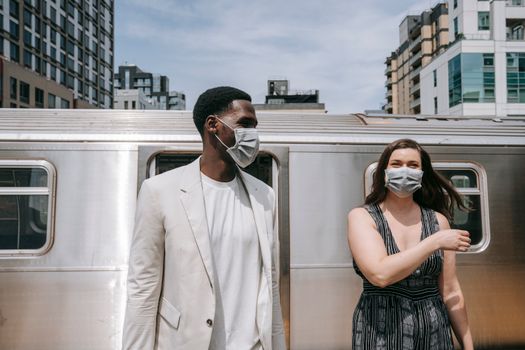 A couple wearing protective face masks standing at a train station, embracing new normal.