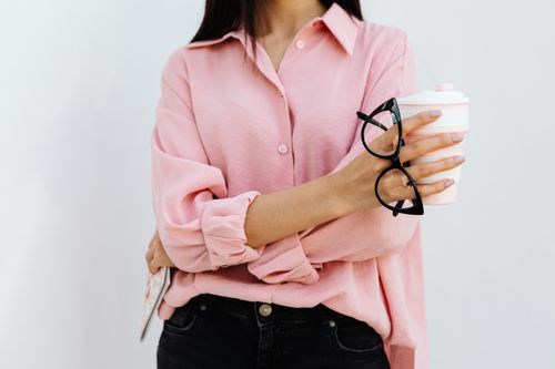 Free Close-up of a stylish woman in a pink shirt holding a coffee cup and eyeglasses, indoors. Stock Photo