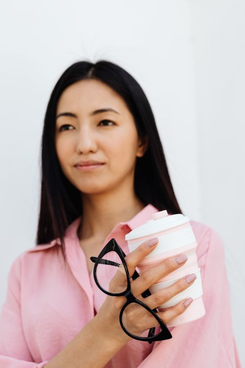 Free Asian woman in pink shirt holding a coffee cup and eyewear, captured in a minimalist style. Stock Photo