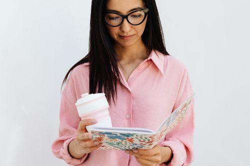 Free Young woman in glasses reading a journal while holding a coffee cup. Stock Photo