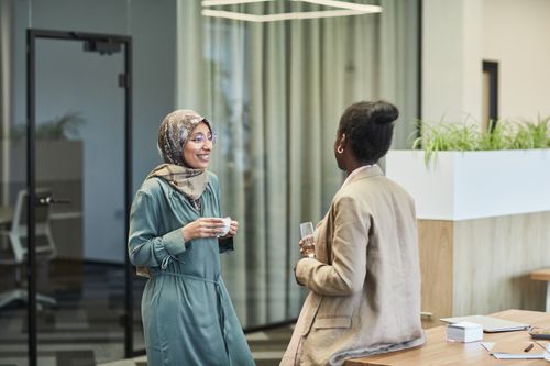 Free Two diverse women colleagues chatting in a modern office setting, fostering workplace communication. Stock Photo