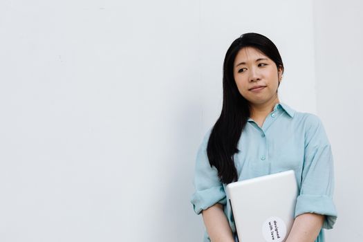 Young Asian woman in a blue shirt holding a laptop with thoughtful expression indoors.
