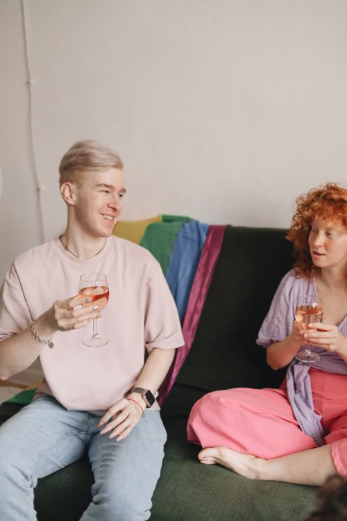 Free Two friends enjoying wine and conversation on a sofa with a rainbow flag backdrop. Stock Photo