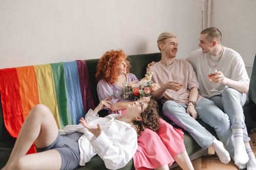 Free A joyful indoor celebration with friends sitting together under a rainbow flag, holding wine and flowers. Stock Photo