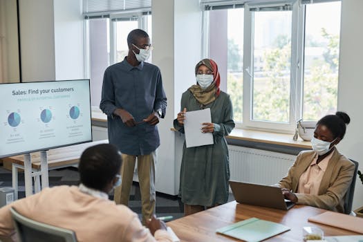 Office meeting with diverse colleagues wearing masks, presenting sales data.