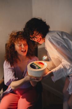 Two women celebrating a birthday with a cake featuring rainbow colors and lit candles.