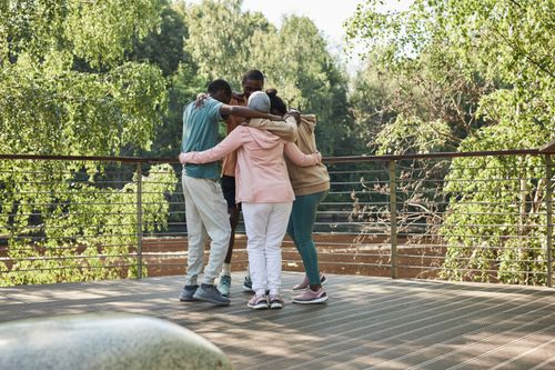 Free Friends huddling in a group hug on a sunny outdoor deck surrounded by greenery. Stock Photo