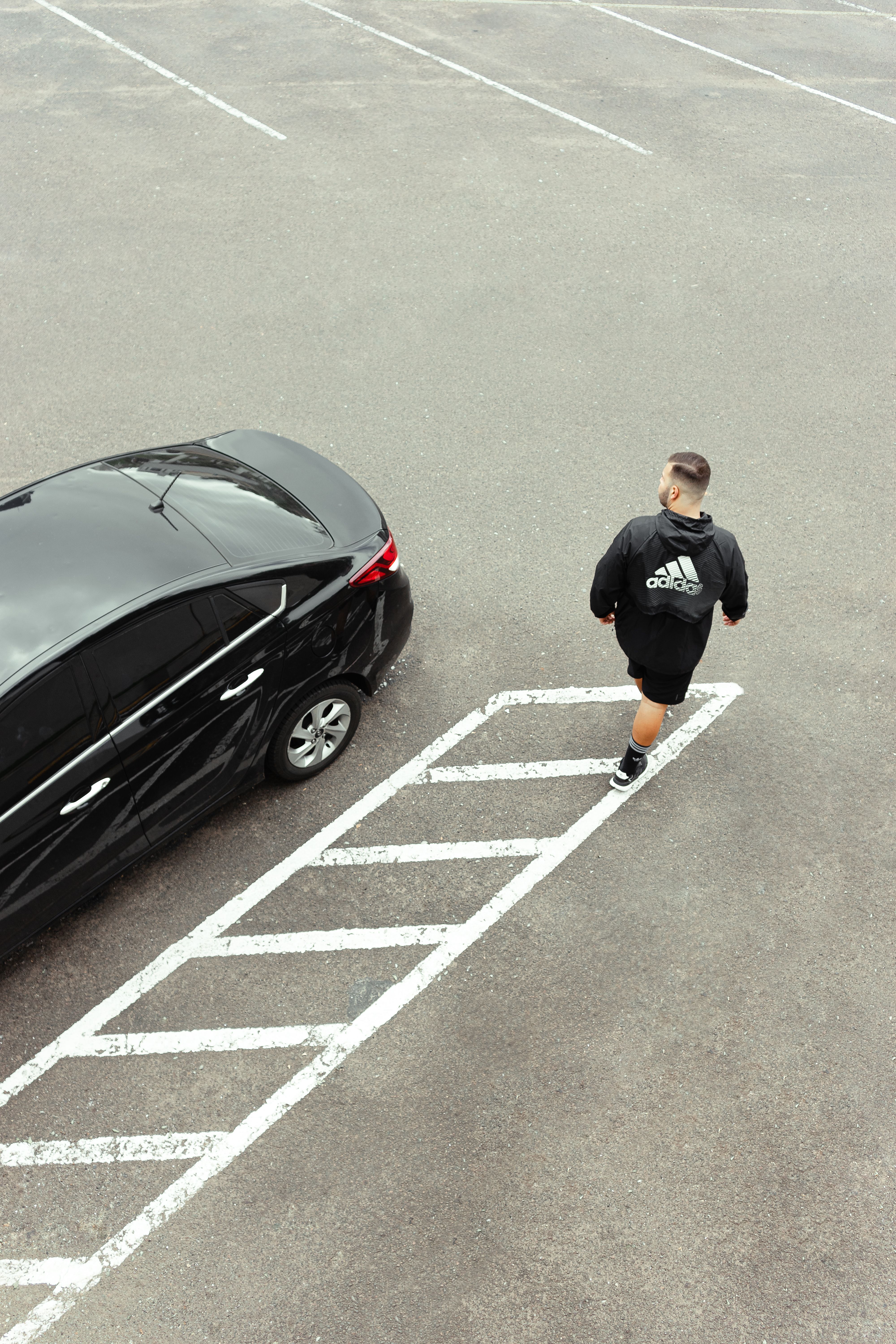 Aerial image of a man in sportswear walking near a parked black car in an empty asphalt lot.