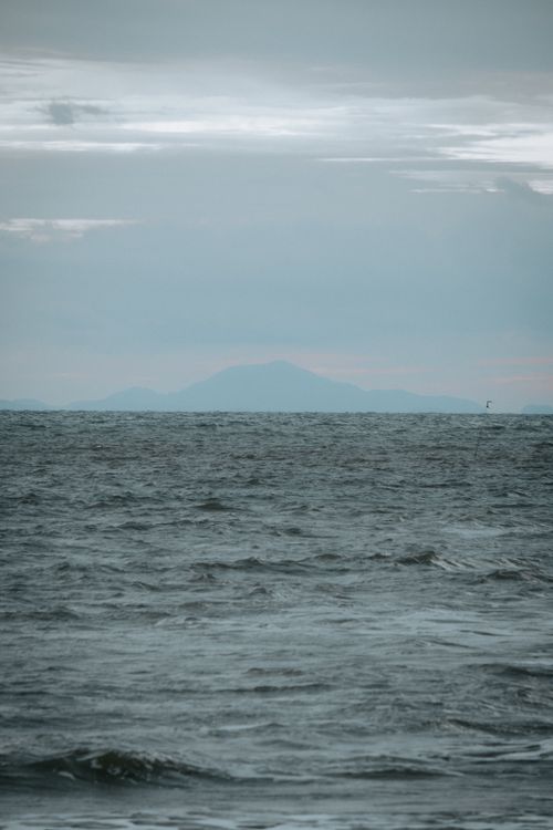 Free Calm ocean waves with a mountain silhouette under a cloudy sky. Stock Photo