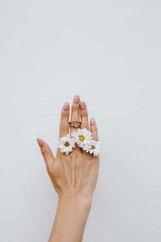 Close-up of a woman's hand adorned with daisy flowers and rings against a white background.