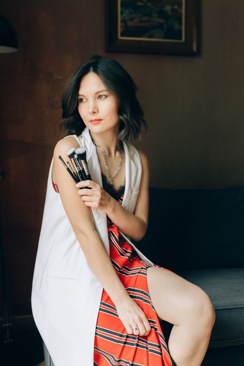 Free A woman in a red dress sits indoors, holding makeup brushes, exuding elegance. Stock Photo