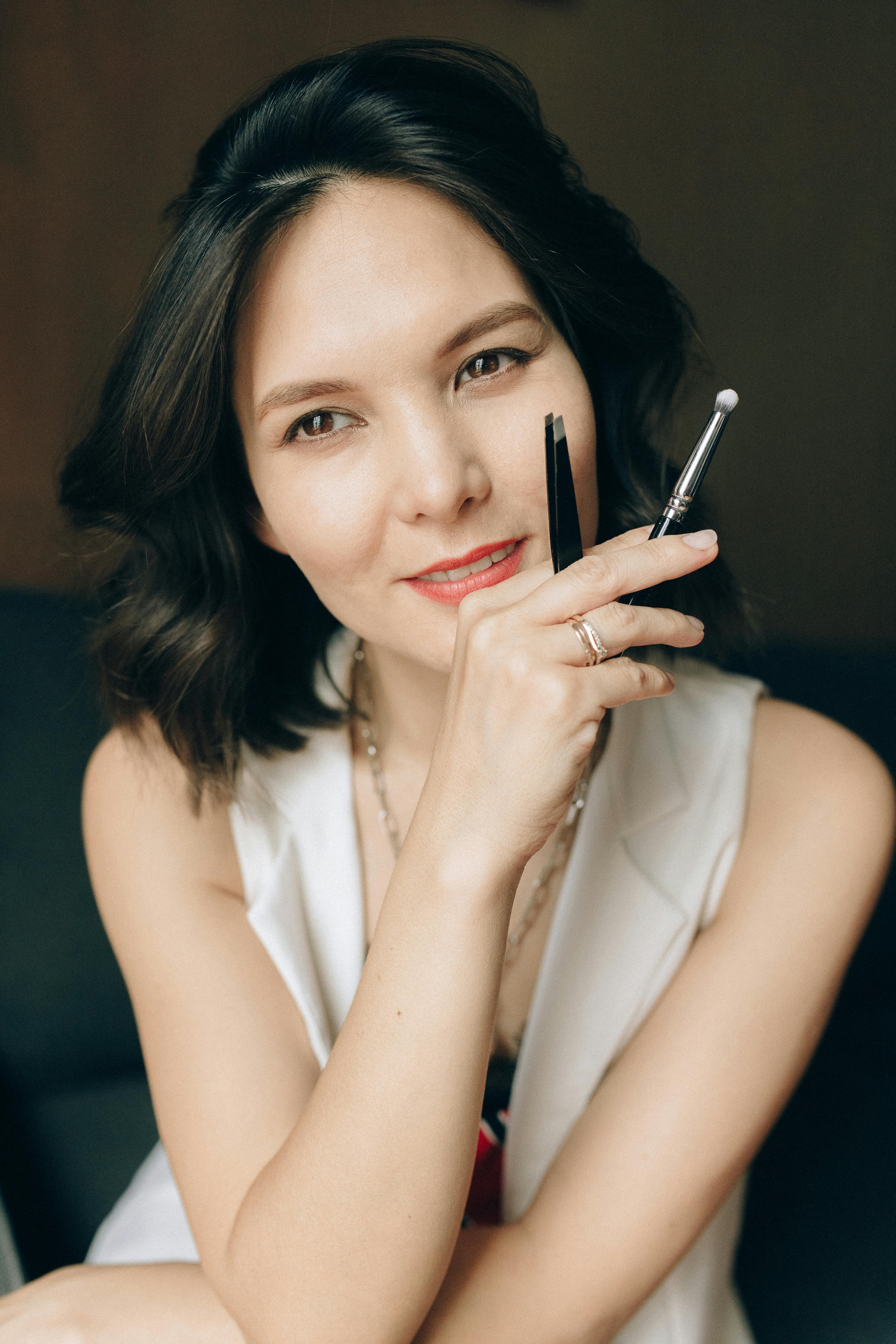 Smiling Asian woman holding makeup brushes, ready for beauty routine.