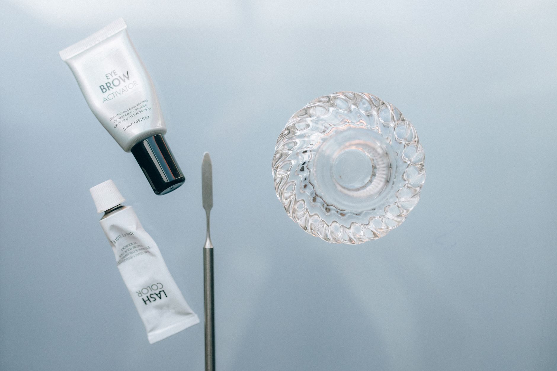 Top view of eyebrow and lash activating cosmetics with a glass dish on a reflective surface.