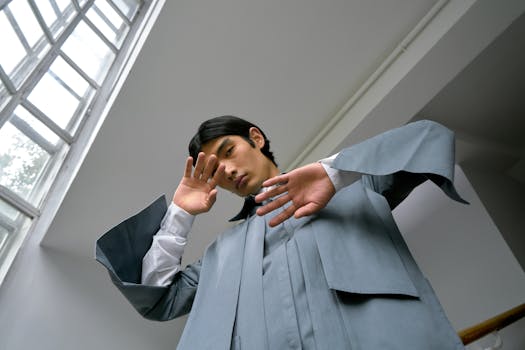 Stylish Asian man posing indoors with a serene expression, wearing modern clothing by a glass window.