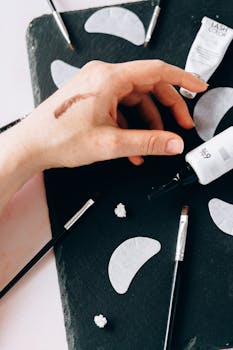 Close-up of cosmetic products and tools on a slate surface, featuring a hand with makeup swatch.