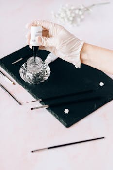 A gloved hand holding a nail polish bottle over a slate with nail art tools.
