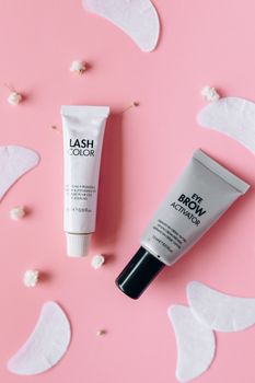 Flat lay of eyelash and eyebrow cosmetic tubes on pink background with floral accents.