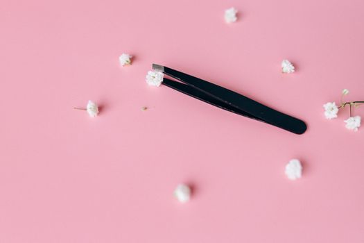 Black tweezers with delicate white flowers on a soft pink background, ideal for beauty themes.
