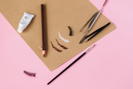 A collection of eyebrow makeup tools and cosmetics arranged on a pink surface for beauty styling.