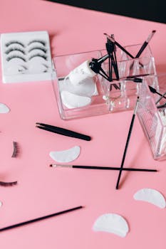 A detailed flat lay of makeup accessories on a pink background featuring false eyelashes and cosmetic brushes.