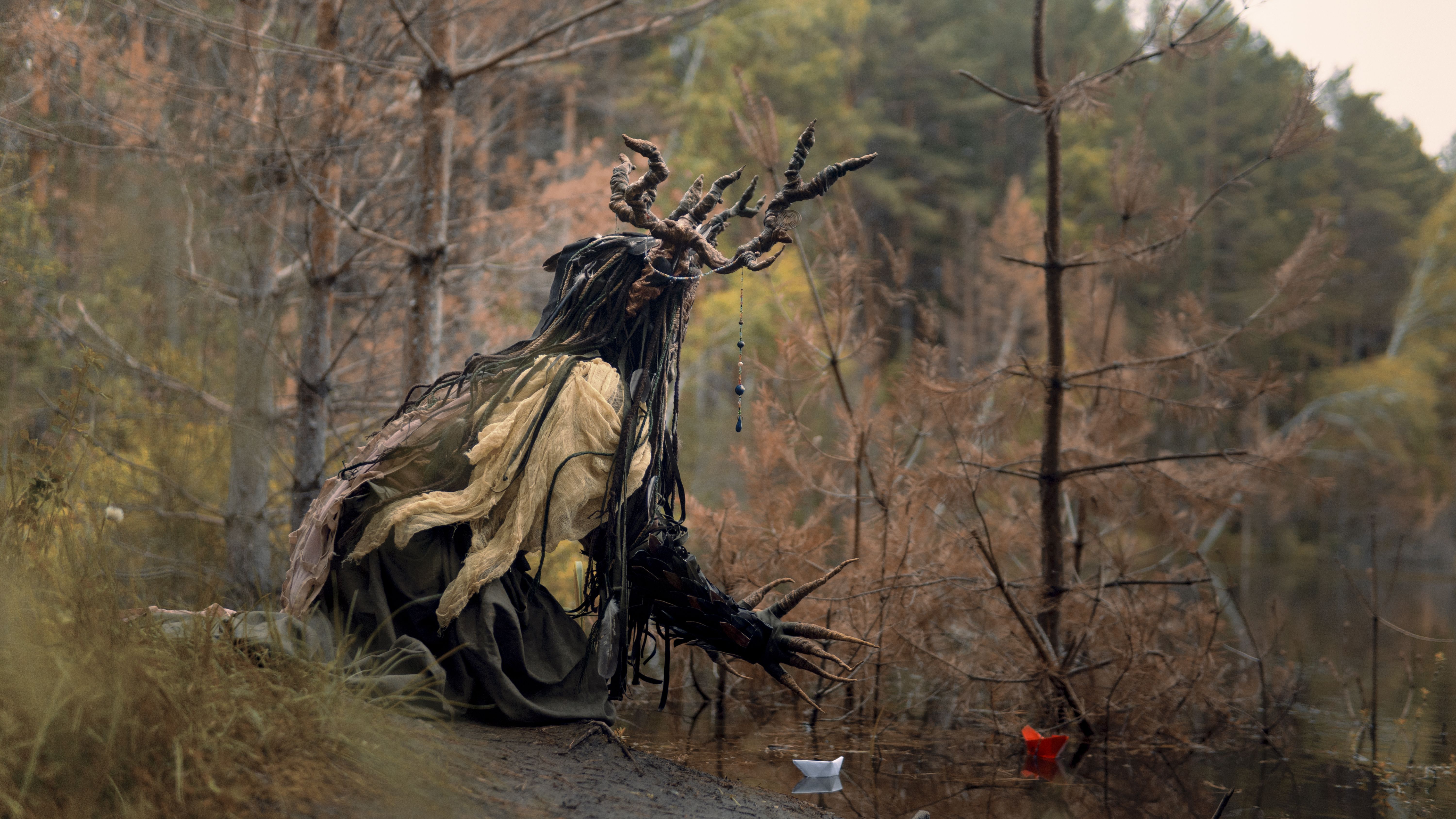 Eerie forest figure with antlered costume sitting by a tranquil lake in autumn.