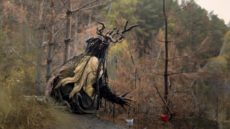 Person In A Frightening Costume With Antlers Crouching On The Lake Shore 