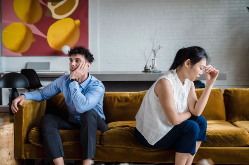 Free A couple sits apart on a sofa, looking unhappy and deep in thought, reflecting relationship conflict. Stock Photo