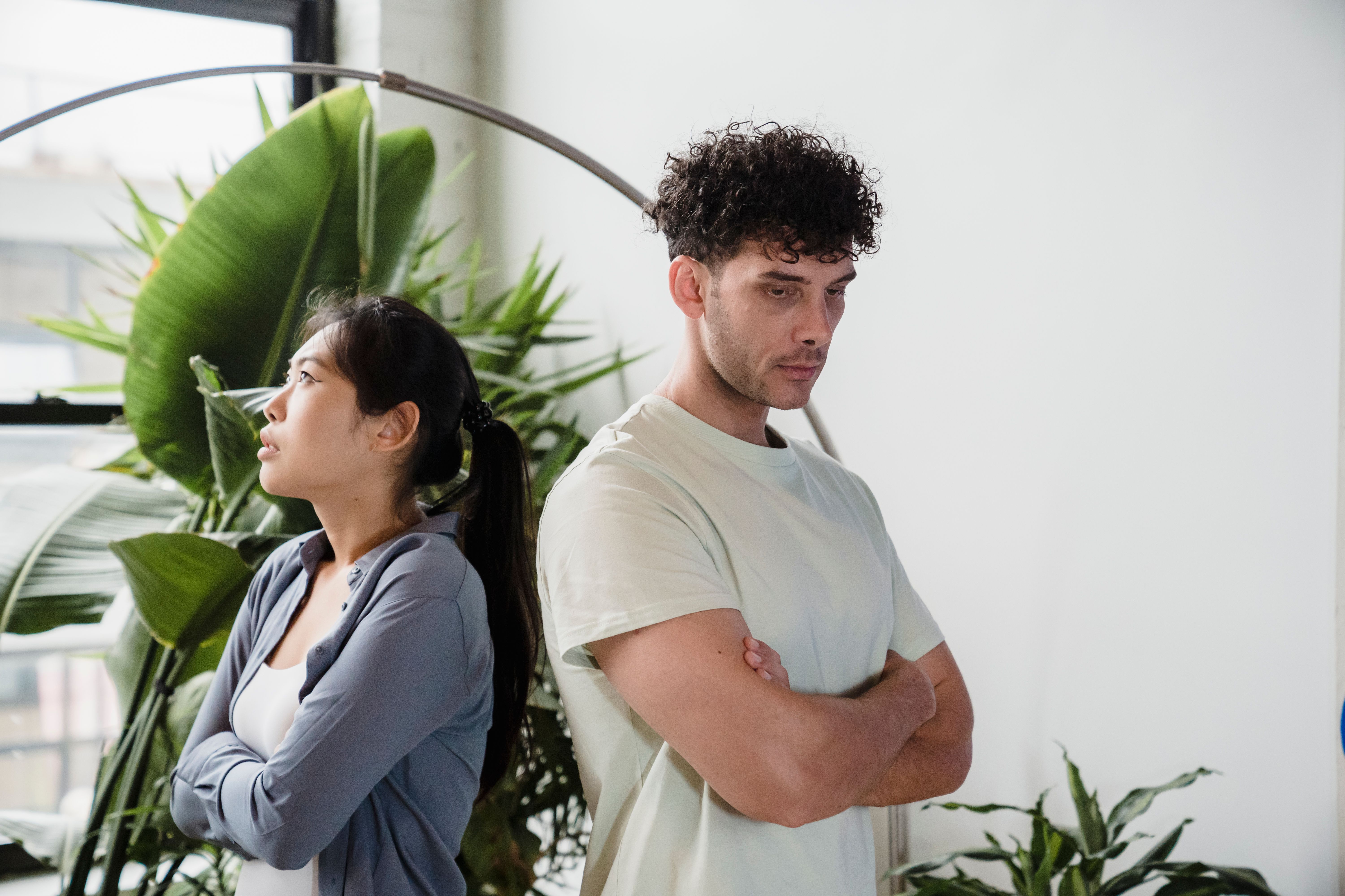 A young couple stands back to back in a room, both appearing upset amidst indoor plants.