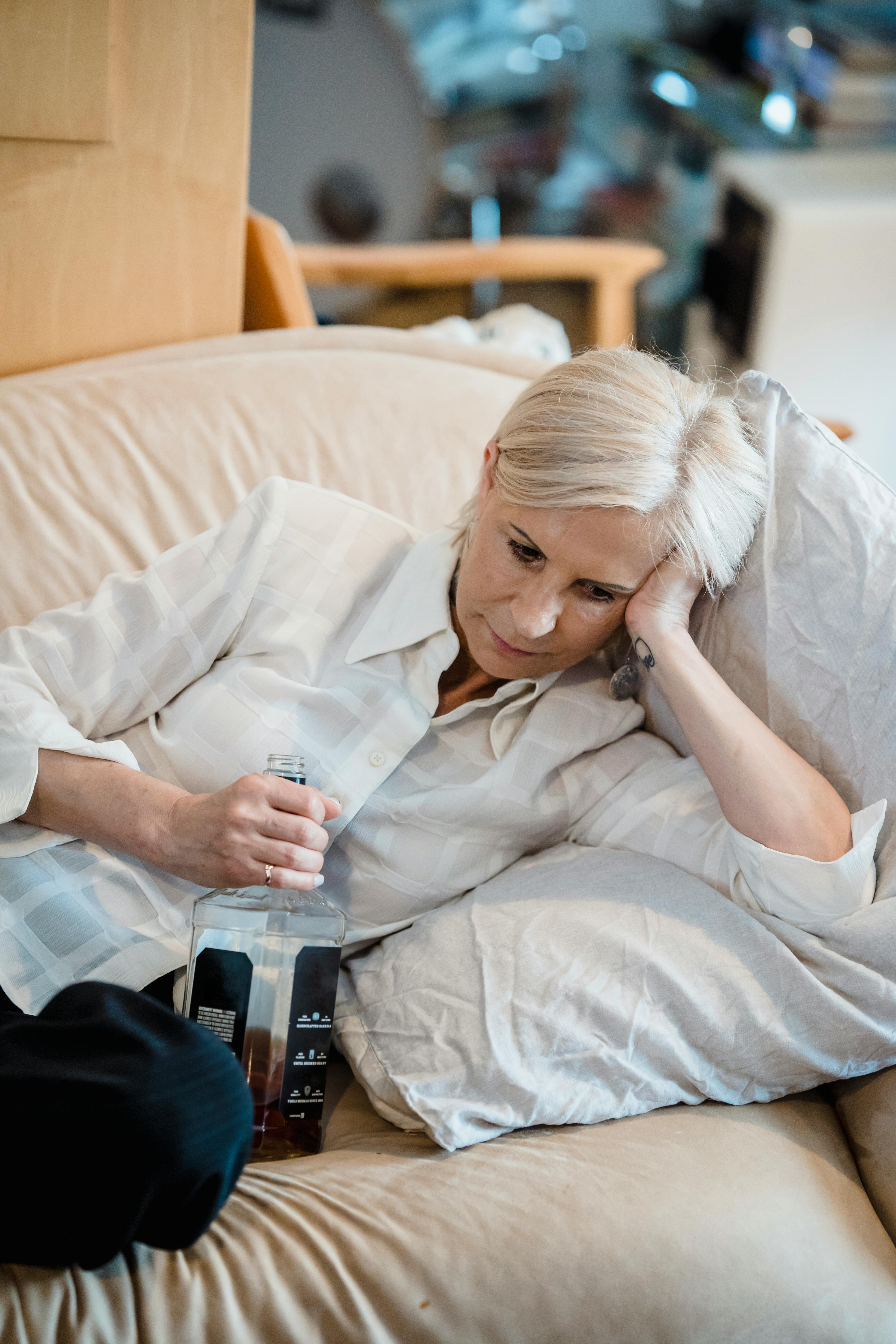 An elderly woman in a white shirt holds a bottle while resting on a sofa indoors.