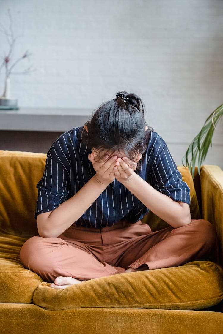 A Frustrated Woman Sitting On A Couch While Covering Her Face Using Her Hands