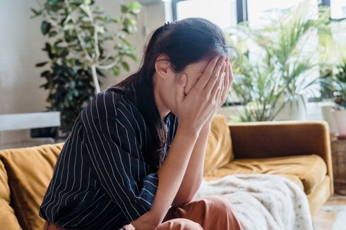 Free Woman covering face with hands, looking frustrated while sitting on a sofa in a modern living room. Stock Photo