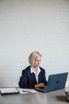 Businesswoman in formal attire typing on laptop at office desk with white background.
