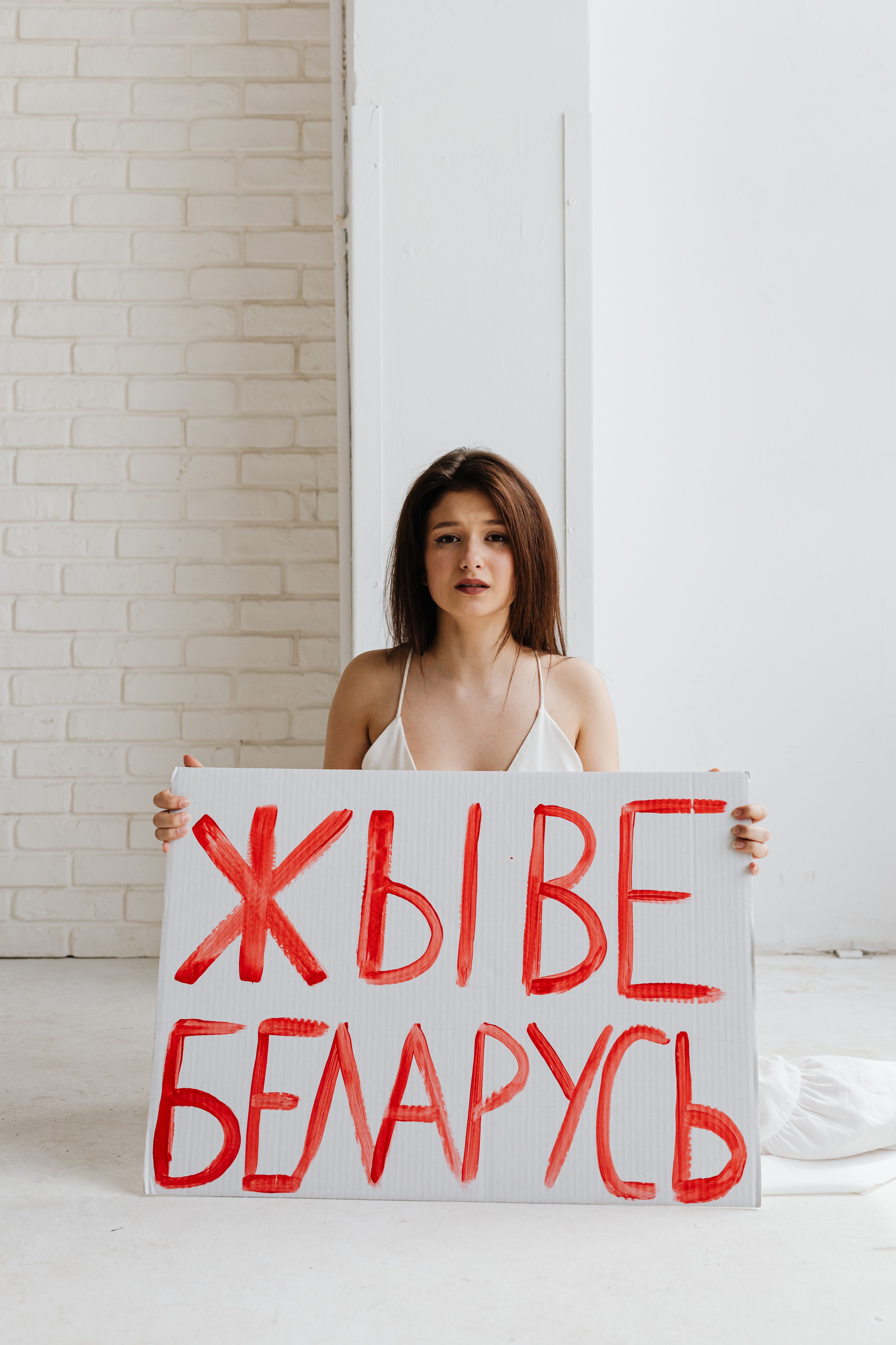 Woman holding a protest sign with Belarusian text in a bright indoor setting.