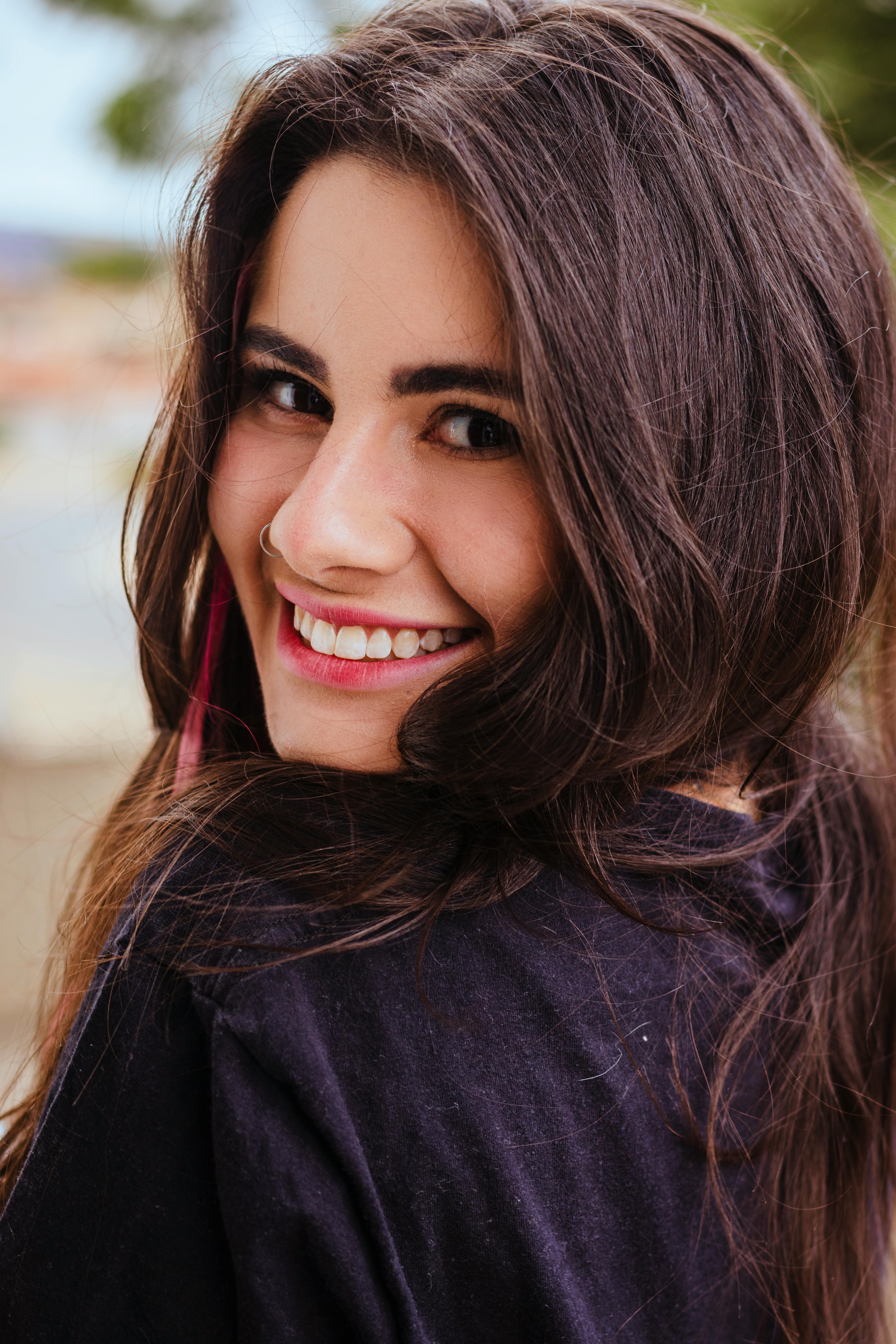 A beautiful woman with long brunette hair smiles warmly at the camera in an outdoor setting.