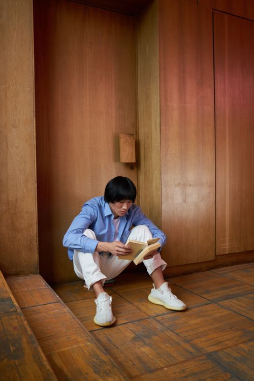 Free Adult man in blue shirt sitting on floor, reading book against wooden wall. Stock Photo