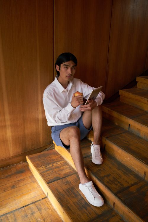 Free Asian man sitting on wooden stairs holding a tablet, wearing white shirt and denim shorts. Stock Photo