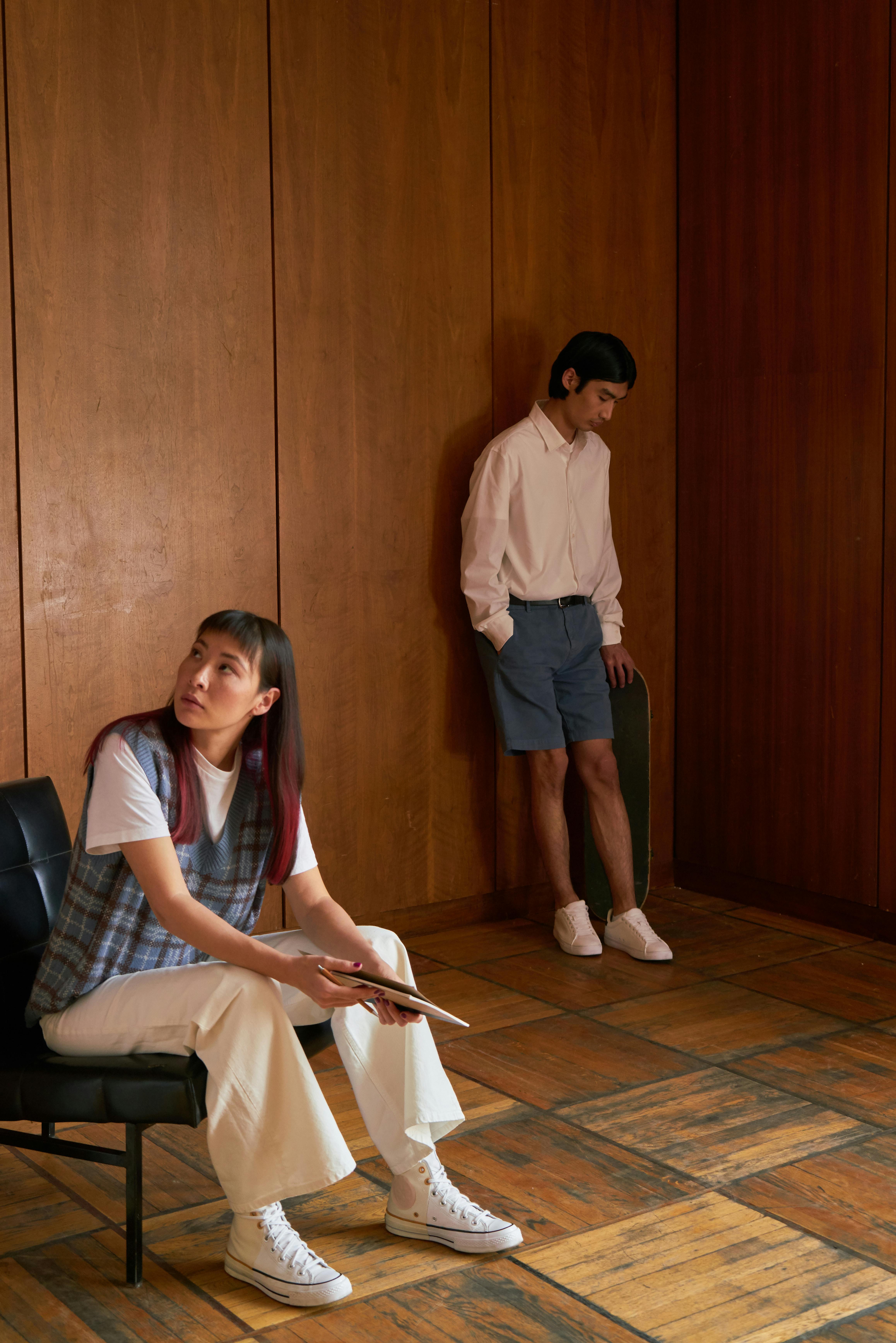 Free Two friends in casual outfits in a wooden room, one seated, one standing. Stock Photo