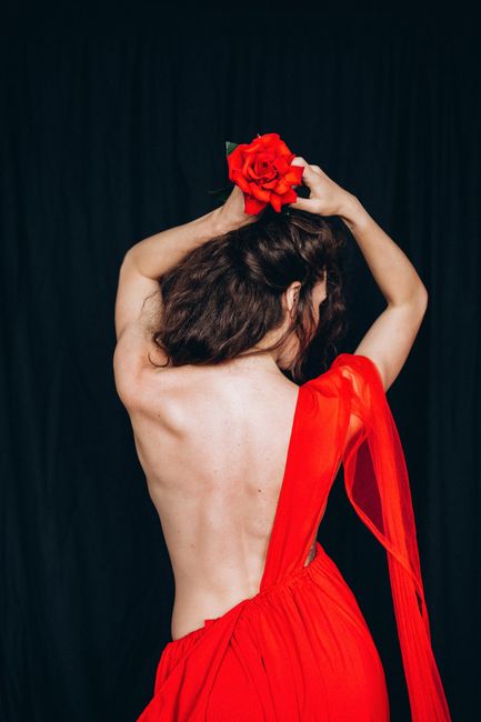 A woman in a red dress poses elegantly with a rose against a dark backdrop.