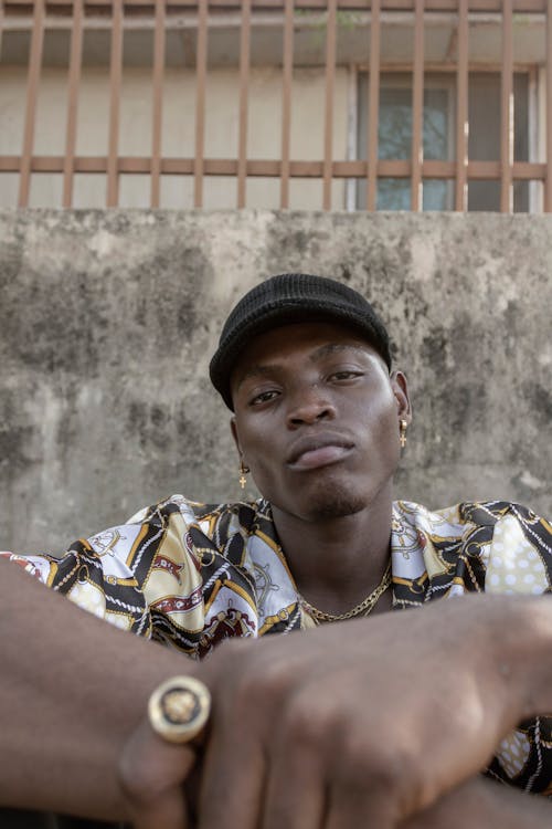 Young African American male in trendy cap and fashionable shirt with ring on finger and stud earrings sitting near metal fence and looking at camera