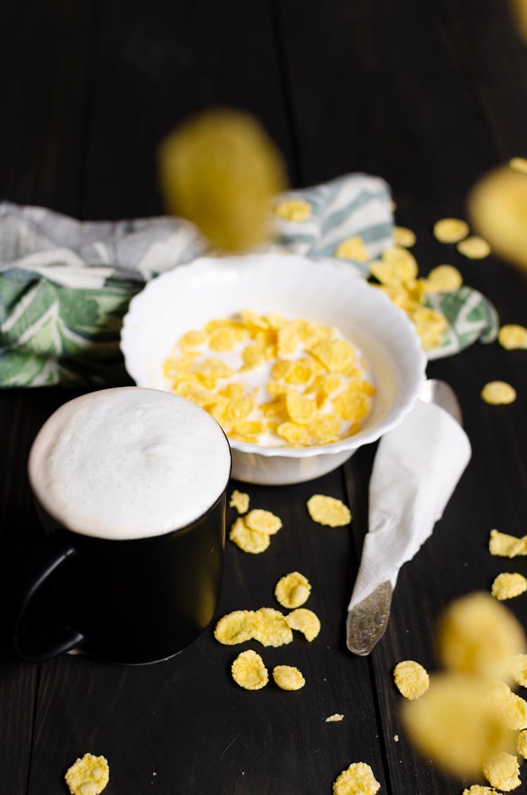 A Mug Of Coffee Beside A Bowl With Corn Flakes On A Black Wooden Surface