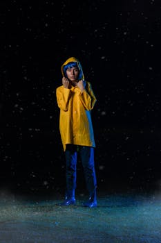 A woman wearing a yellow raincoat stands on grassy ground in rain at night.