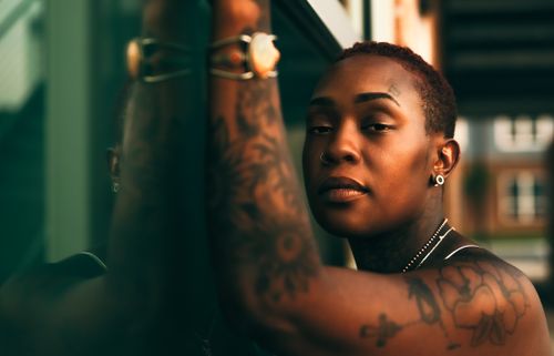 Free Portrait of a tattooed woman with short hair leaning against a window, exuding confidence. Stock Photo