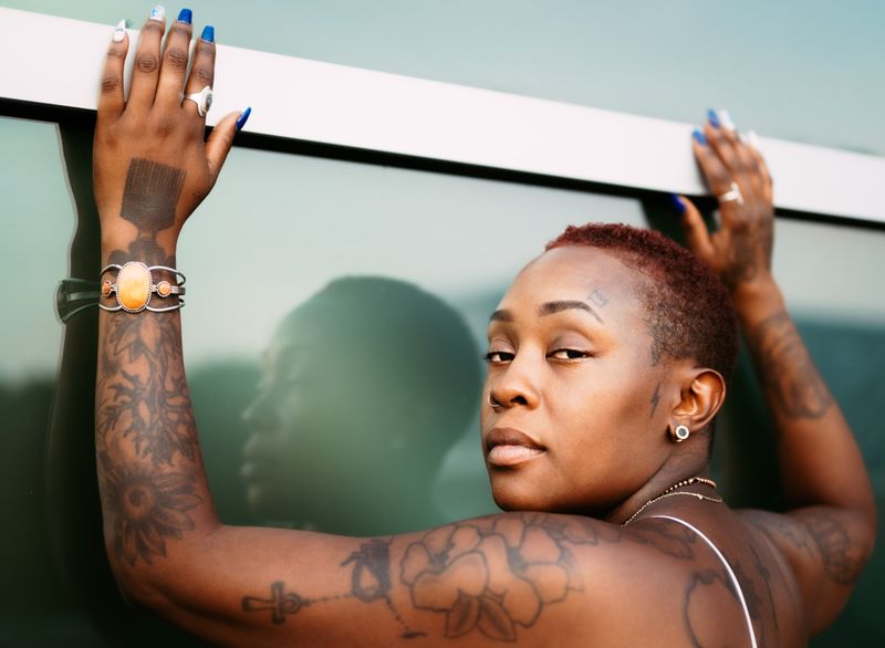 A Black woman with fine-line tattoos across her arms, shoulder and hands, wearing a nose stud and an ear stud.