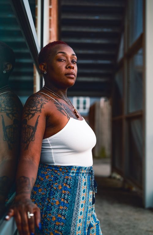 Free Tattooed woman in a white top poses confidently outdoors, leaning against a glass wall. Stock Photo