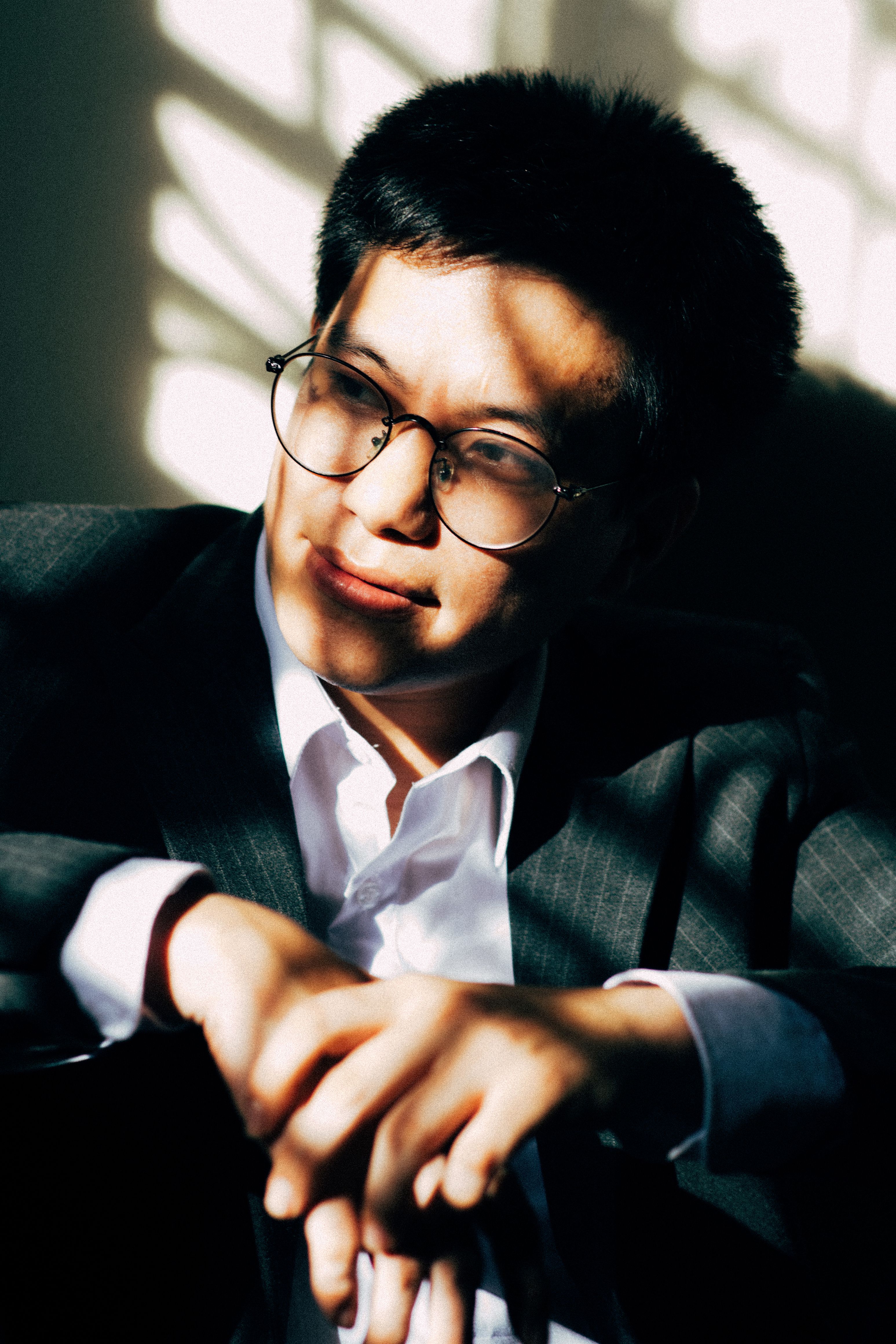 Asian man in a suit, captured in shadows, wearing eyeglasses and posing thoughtfully indoors.