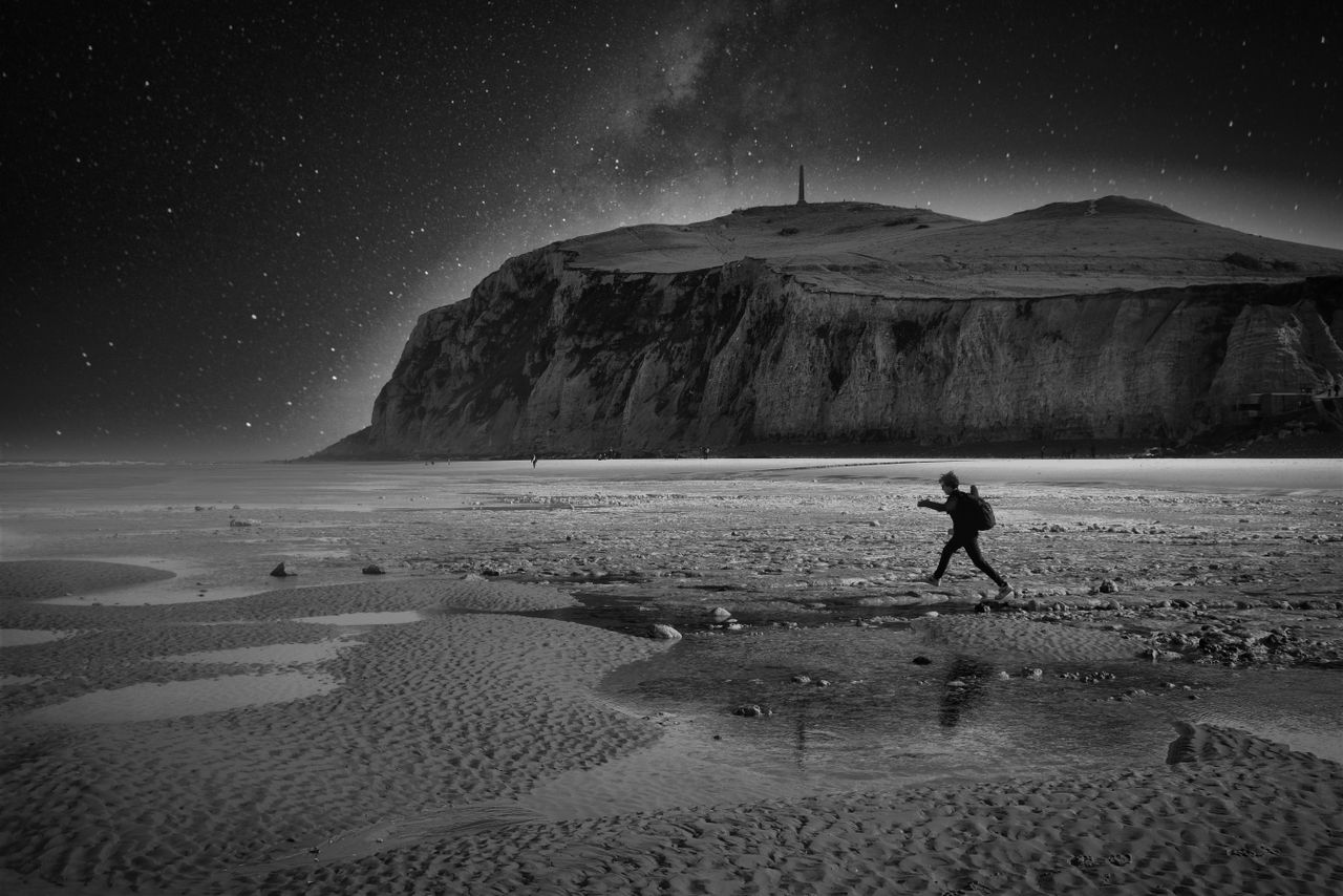 a person walks across a rocky beach under a starry sky by a dramatic c