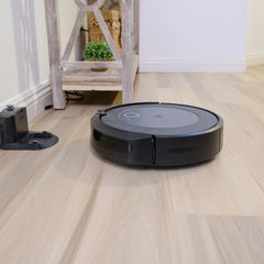 A dark robot vacuum on a pale wooden floor with its dock by the wall
