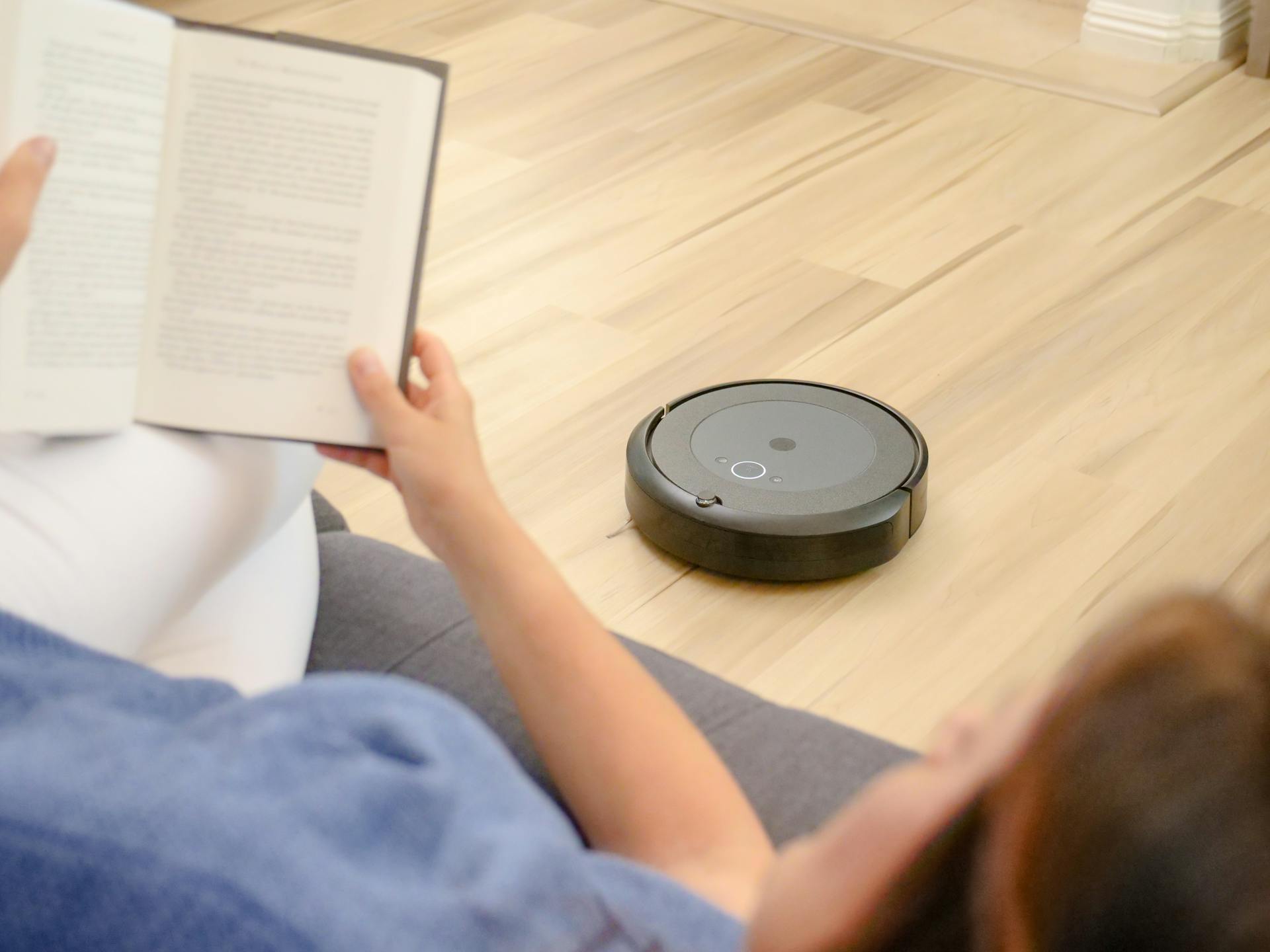 A woman relaxes on a sofa reading while a robotic vacuum navigates the floor.